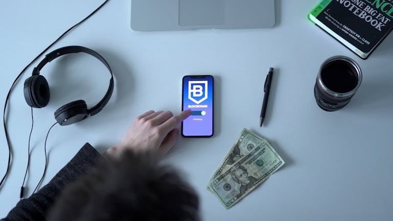 Overhead view of blockchain app on smartphone with cash, headphones, and notebook.